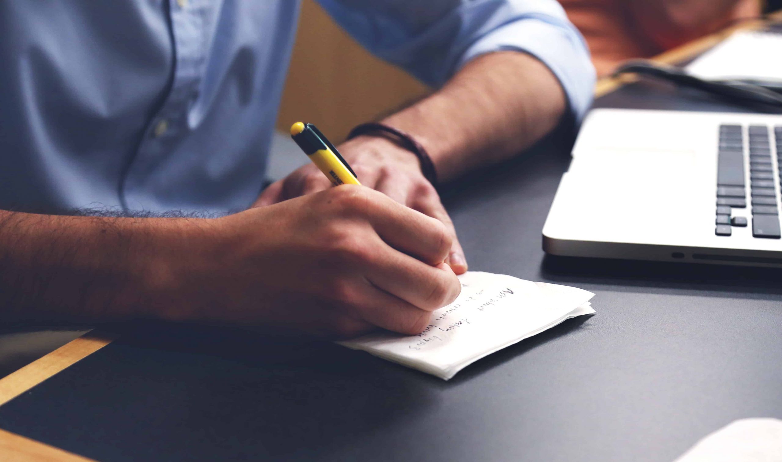 Person writing notes on a notepad beside an open laptop at a desk
