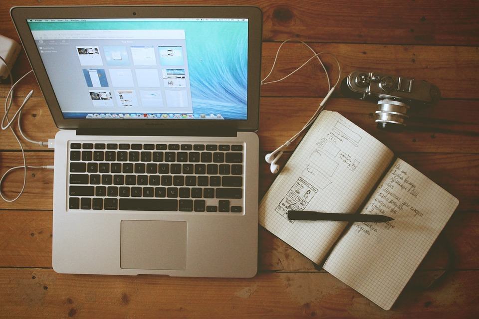 MacBook, open notebook with wireframe sketches, earphones, and a vintage camera on a wooden desk