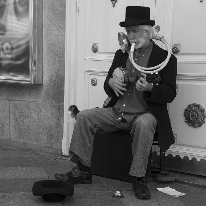 Street musician sitting outside a building playing a ukulele with a horn attached, illustrating a one-man-band