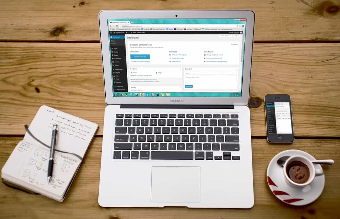 MacBook Air showing the WordPress dashboard on a wooden desk with a coffee cup and notebook